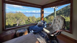 A room with a view of a lake and mountains, a wicker chair, a small table, and two ottomans. - Tata Beach Heights - Spectacular Ocean and Bush Views (Tata Beach)