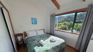 A bedroom with a bed, a chair, a nightstand, a window with curtains, and a view of greenery outside. - Tata Beach Heights - Spectacular Ocean and Bush Views (Tata Beach)
