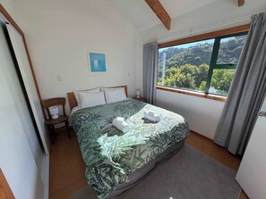 A bedroom with a bed, a chair, a nightstand, a window with curtains, and a view of greenery outside. - Tata Beach Heights - Spectacular Ocean and Bush Views (Tata Beach)