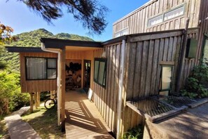A wooden cabin with a covered porch, a bicycle, and a view of the surrounding forest. - Tata Beach Heights - Spectacular Ocean and Bush Views (Tata Beach)