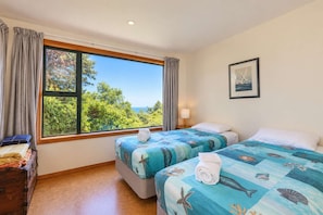 A hotel room with two beds, a window with curtains, a wooden dresser, a framed picture on the wall, and a view of greenery and the sea. - Tata Beach Heights - Spectacular Ocean and Bush Views (Tata Beach)
