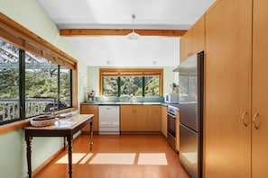 A kitchen with wooden cabinets, a stainless steel refrigerator, a built-in oven, and a window overlooking a forest. - Tata Beach Heights - Spectacular Ocean and Bush Views (Tata Beach)
