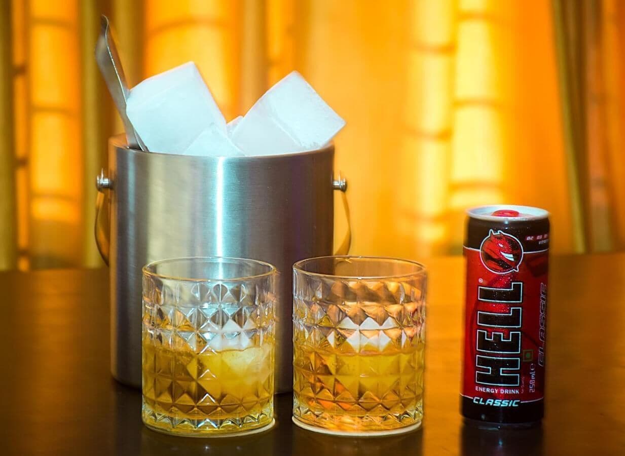 Two glasses of amber liquid, a metal container with a spoon, and a can of energy drink.
