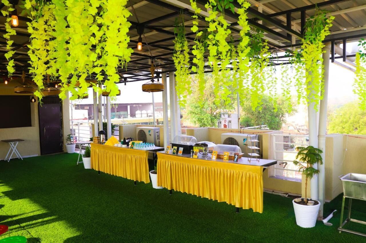 Un espacio para eventos al aire libre con césped artificial verde, manteles amarillos, plantas colgantes y vistas de edificios al fondo.