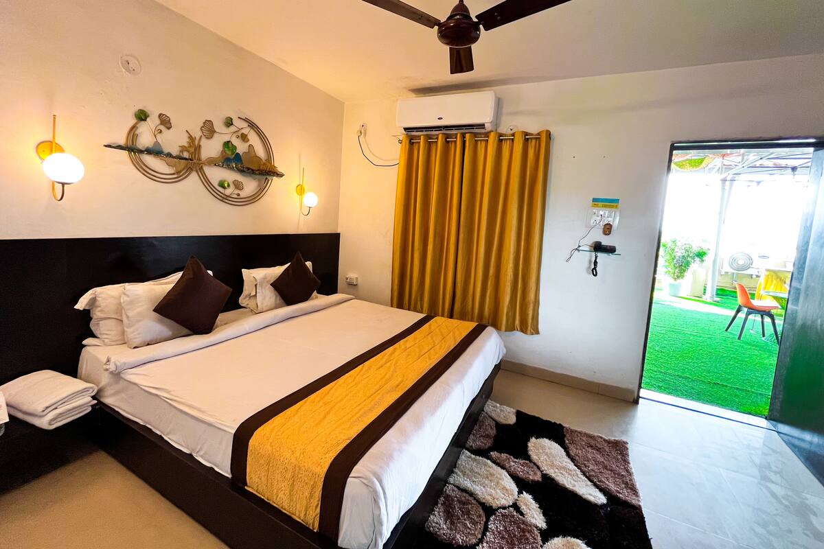 A hotel room with a bed, a ceiling fan, a wall-mounted air conditioner, and a view of a green outdoor area.
