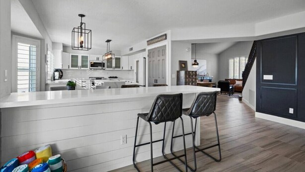 A modern kitchen with white cabinetry, a central island, bar stools, and a dining area with a staircase. - 6th Tee Luxury | Pool & Fire Pit (Chandler)