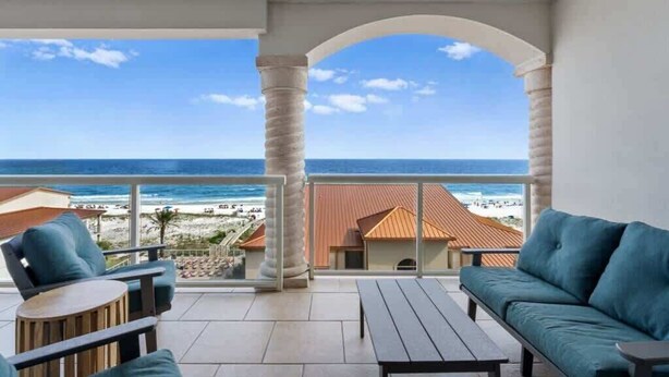 A balcony with a view of the beach and ocean, featuring a sofa, chairs, and a table.