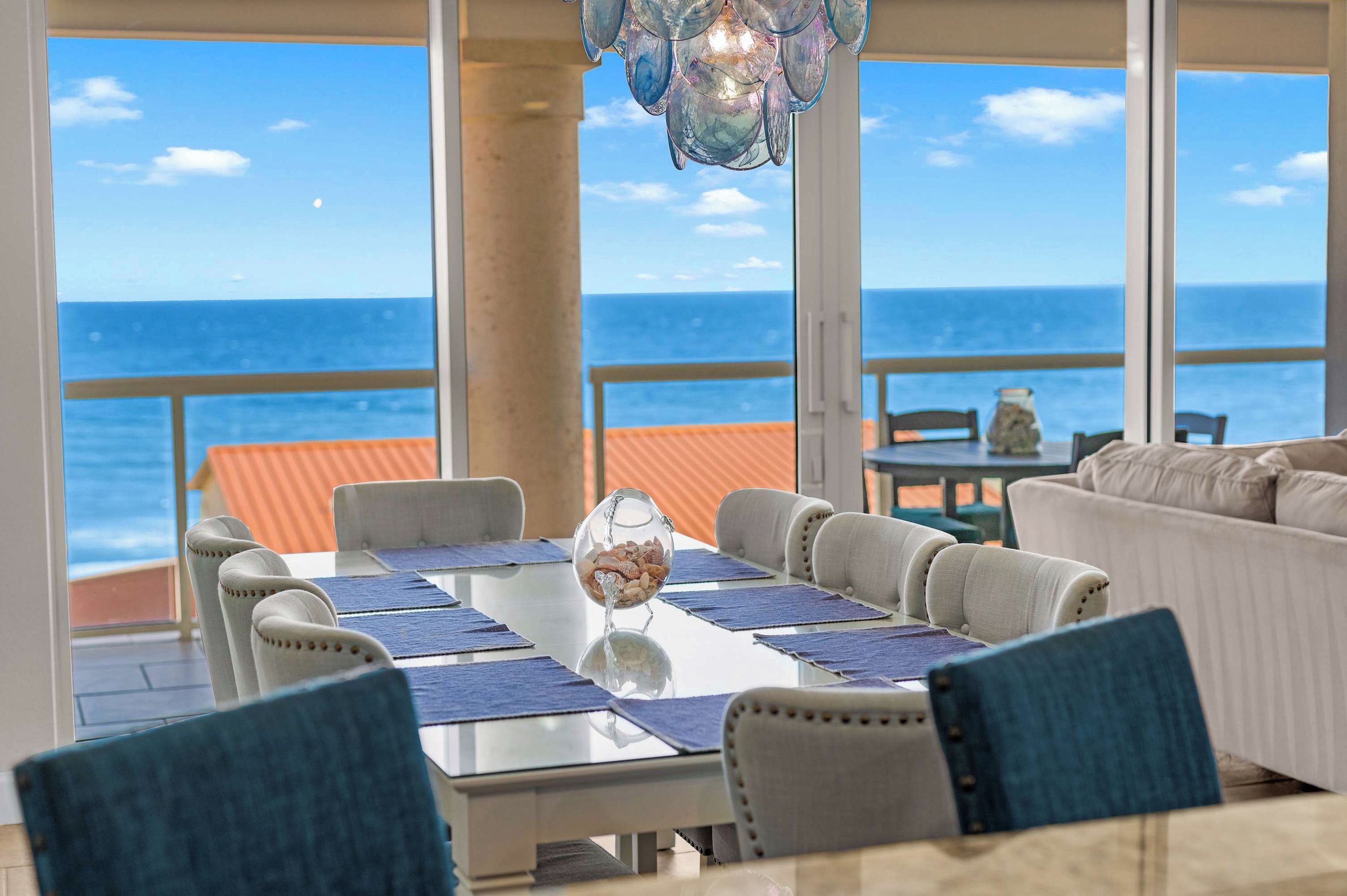 Área de refei ções com vista para o oceano, mesa de vidro com jogos americanos azuis e luminária pendente.