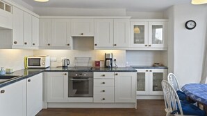 A compact kitchen with white cabinets, a built-in oven, a microwave, and a coffee maker. There is a dining table with chairs and a clock on the wall. - Apartment, Sea Views & Terrace on Waters Edge (Fowey)