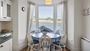 A dining area with a round table and chairs, a window with a view of the sea, and a clock on the wall. - Apartment, Sea Views & Terrace on Waters Edge (Fowey)