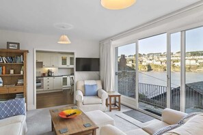 A spacious living room with a sofa, a coffee table, and a bookshelf. A kitchen is visible in the background, and a balcony with a view of the city is also present.