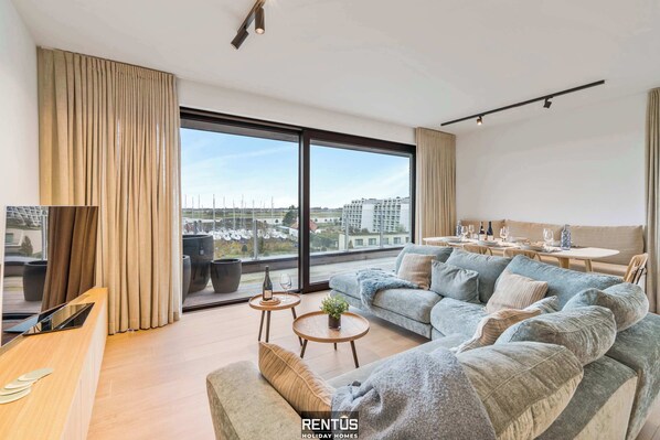 Een moderne woonkamer met een bank, salontafel en televisie. Er is een groot raam met gordijnen, een eethoek met een tafel en stoelen, en een uitzicht op gebouwen.