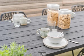 Een houten tafel gedekt met glazen, een mok met dessin, een kom met een lepel en twee bakjes granola.