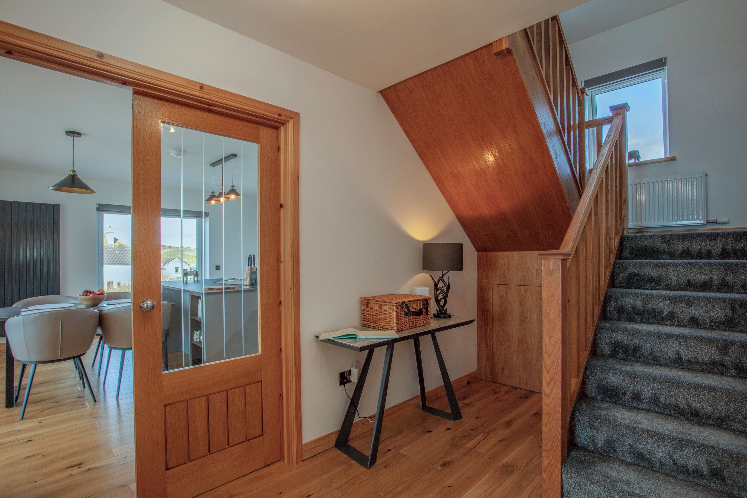 A modern interior with a wooden staircase, a dining area with chairs, and a table with a lamp and basket.