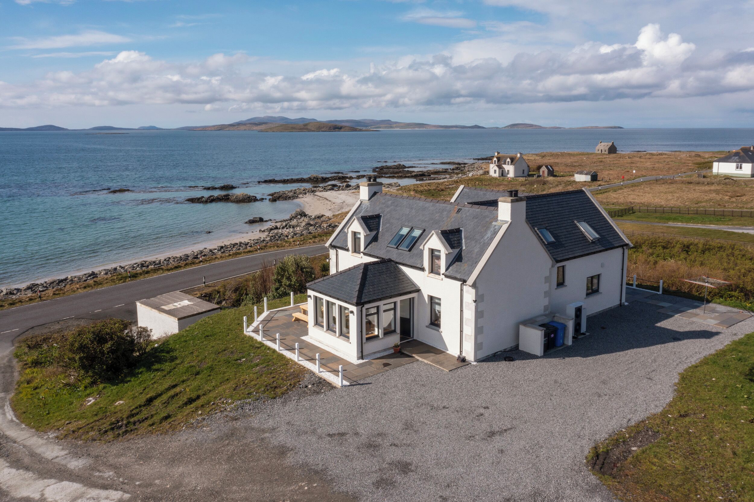 A coastal house with a gravel driveway, a small porch, and a view of the sea and distant hills.