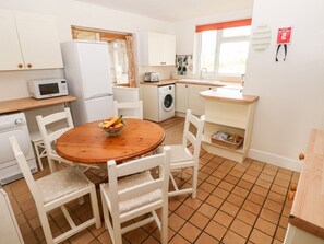 A compact kitchen with white cabinets, a microwave, a washing machine, and a dining area with a wooden table and white chairs.