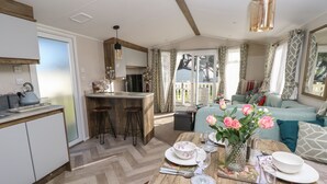 A modern kitchen with a central island, a dining area with a wooden table set for four, and a cozy living area with a sofa and a large window. - Bay View (Bembridge)