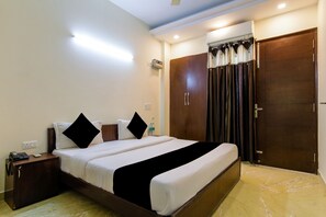 Quarto de hotel com uma cama grande, uma mesinha de cabeceira com telefone e uma garrafa, um guarda-roupa e uma cortina.