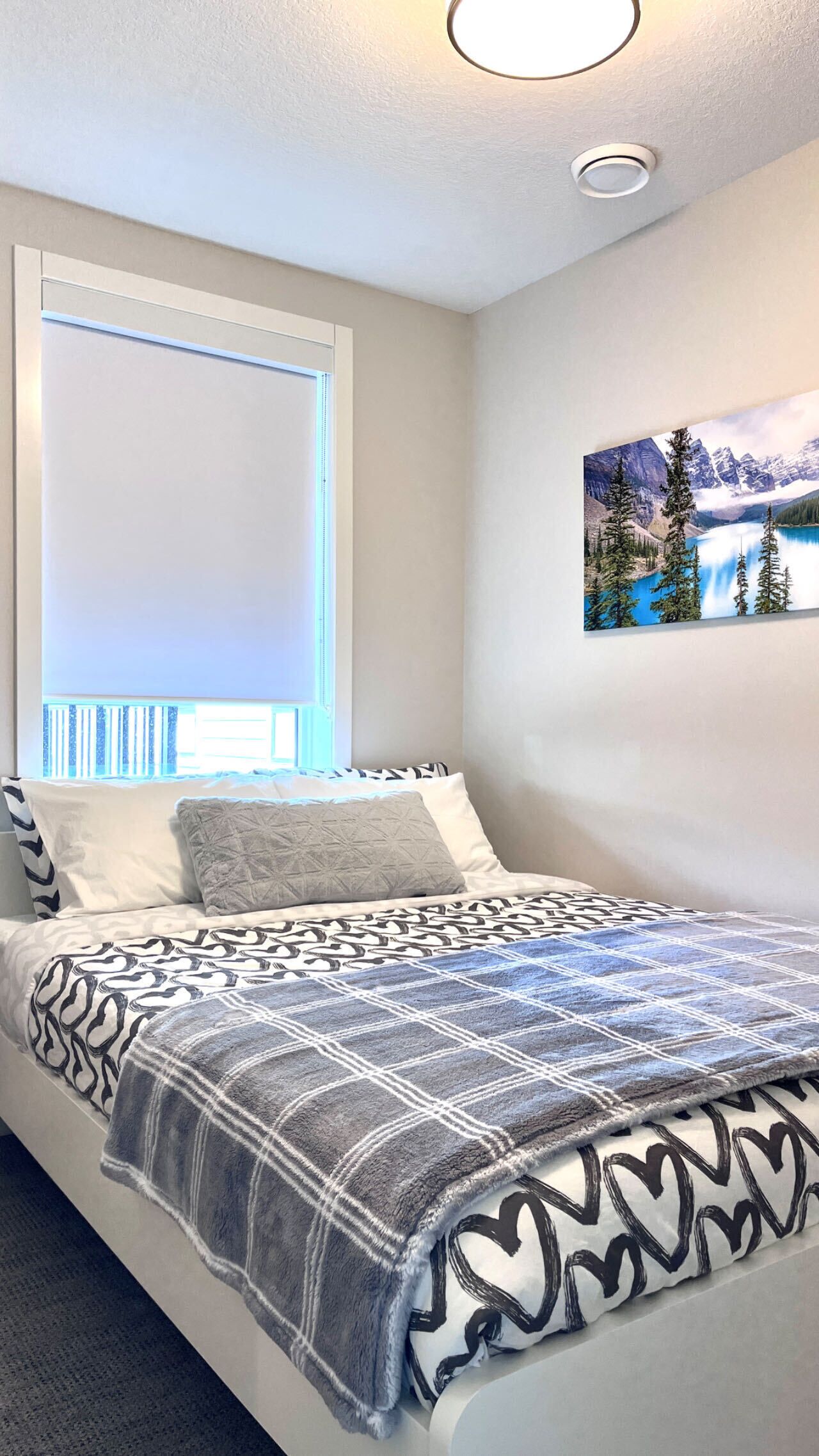Une chambre à coucher avec un lit, une fenêtre avec des stores, un luminaire au plafond et un tableau accroché au mur.
