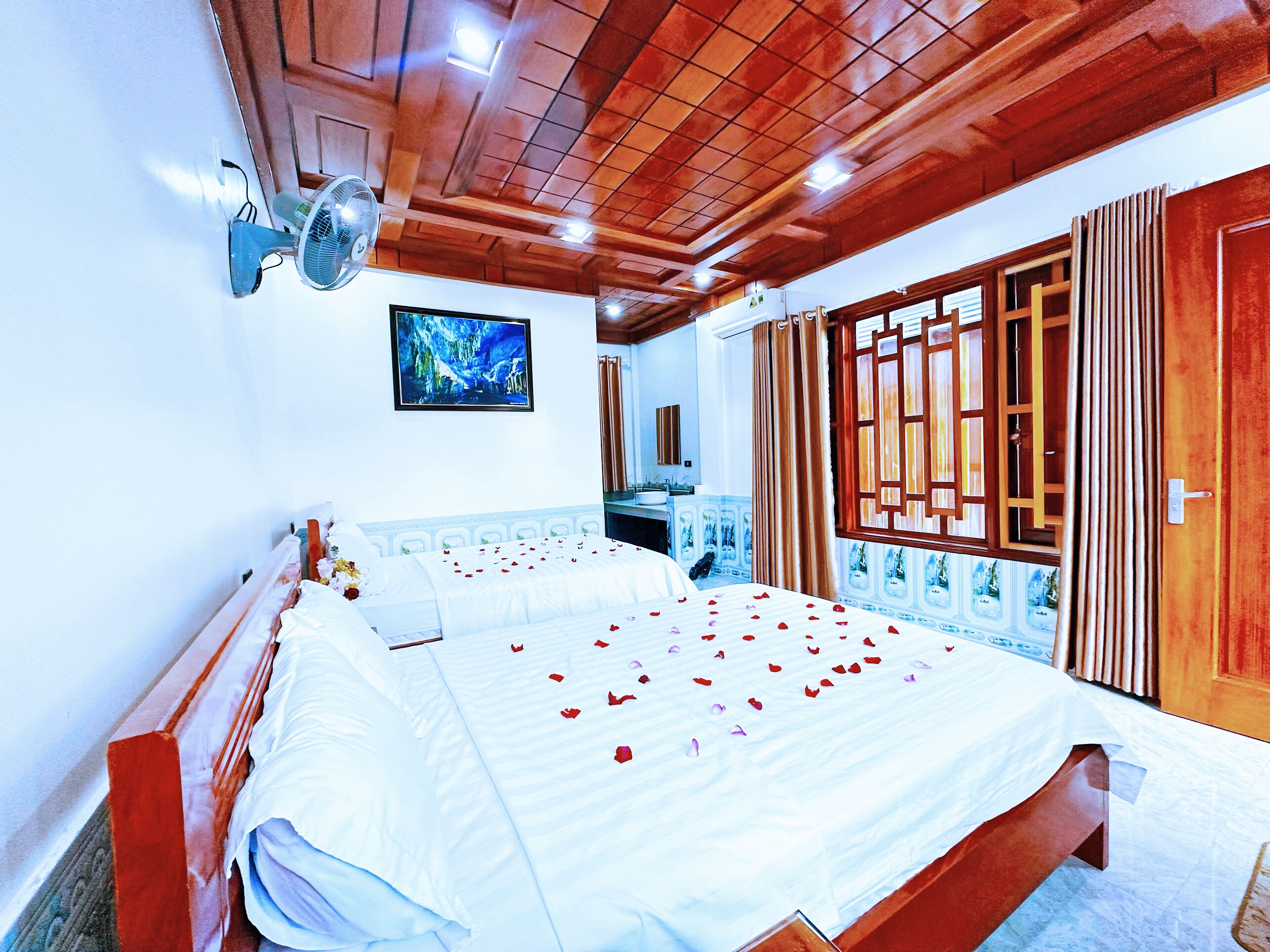 Habitación de hotel con una cama grande, un ventilador de techo, un baño con lavamanos y espejo, y una ventana con cortinas.