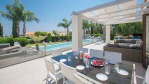 Un espace extérieur aménagé pour les repas, avec vue sur la piscine, des meubles en osier blancs et un coin salon abrité.