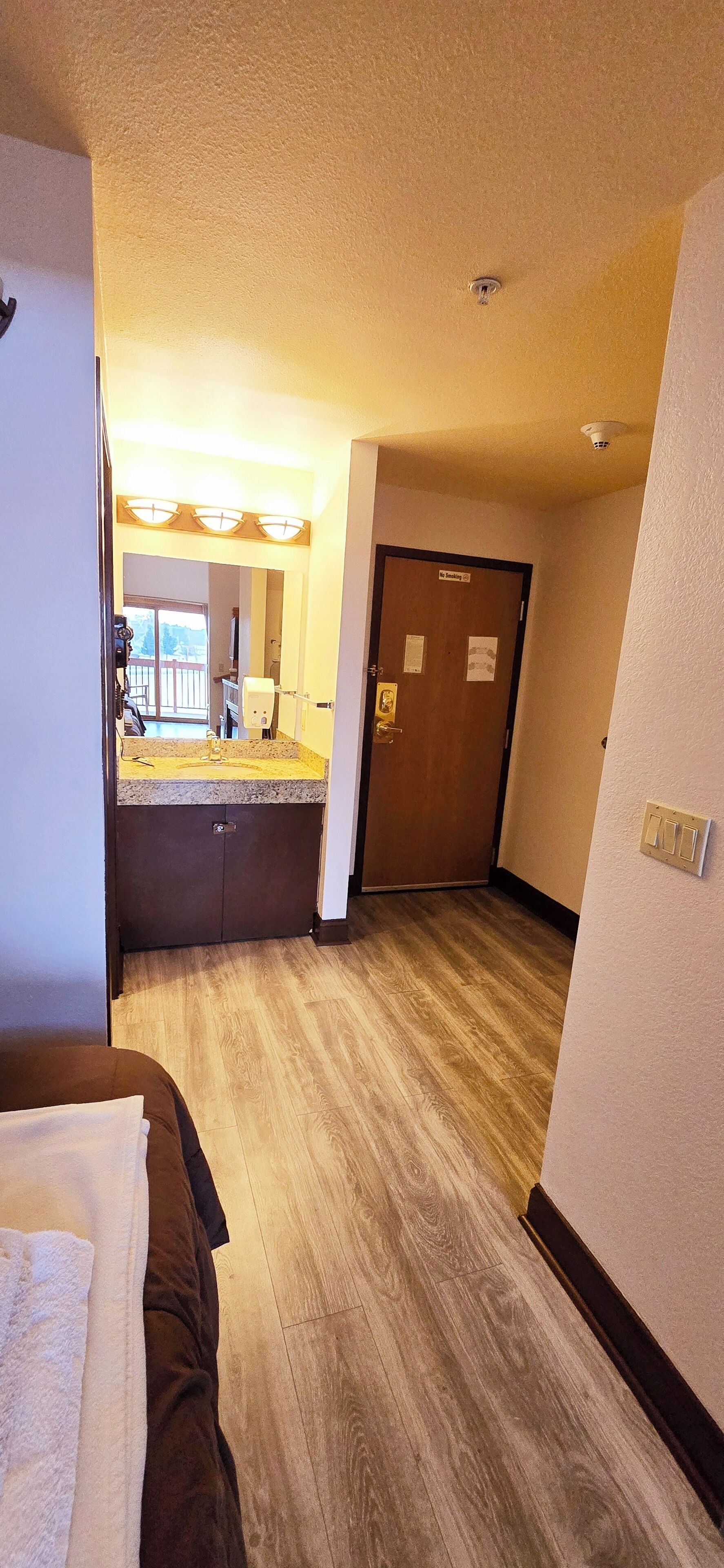 Ein Hotelzimmer mit Holzboden, einem Bett mit weißer und brauner Bettdecke, einem Badezimmer mit Waschbecken und Spiegel sowie einer Tür mit der Aufschrift 'The Lobby'.