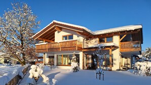 A two-story chalet with a wooden balcony, large windows, and a snow-covered roof.