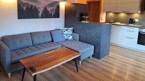 A modern living room with a grey sofa, a wooden coffee table, and a painting of a forest. A kitchen area is visible with white cabinets and a coffee machine.