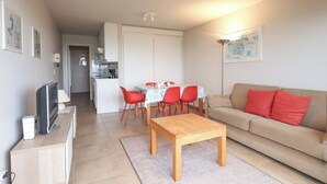 A modern living room with a wooden coffee table, a beige sofa with red cushions, a dining area with a table and chairs, and a kitchen in the background.