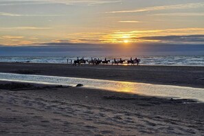 Ein Strand bei Sonnenuntergang, mit Reitern und ruhiger See.