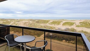 A balcony with a table and chairs, overlooking a coastal landscape with dunes and a road.