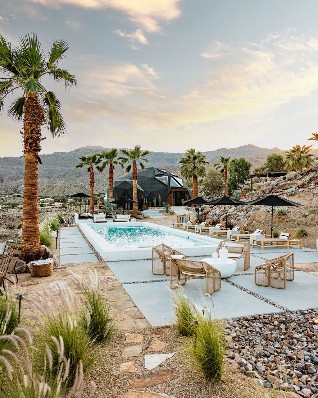 A pool area with lounge chairs and umbrellas, surrounded by palm trees and a mountain backdrop.
