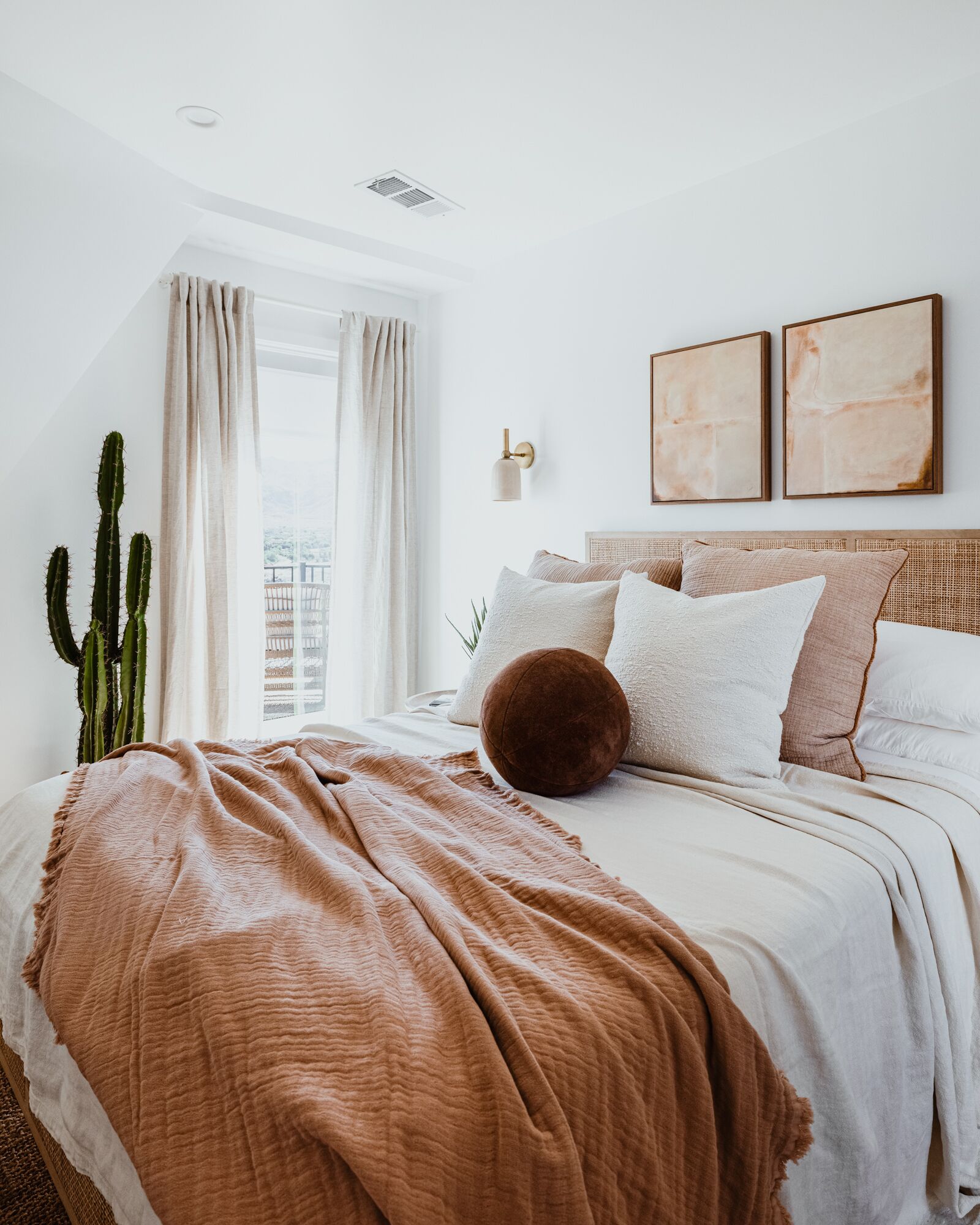 Une chambre avec un grand lit, une fenêtre avec des rideaux, un cactus et deux tableaux abstraits accrochés au mur.