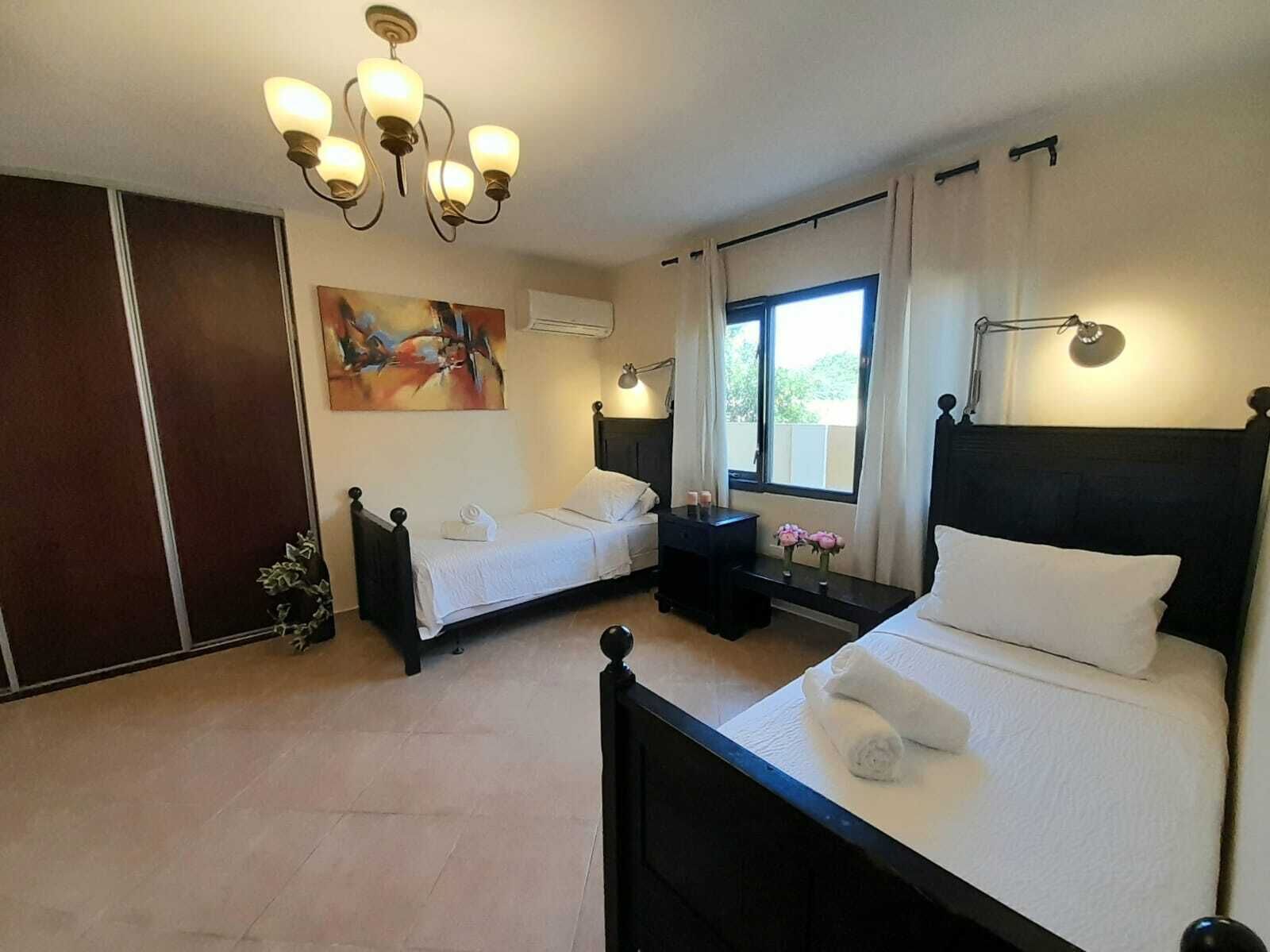 A hotel room with two single beds, a large painting on the wall, a window with curtains, and a chandelier.