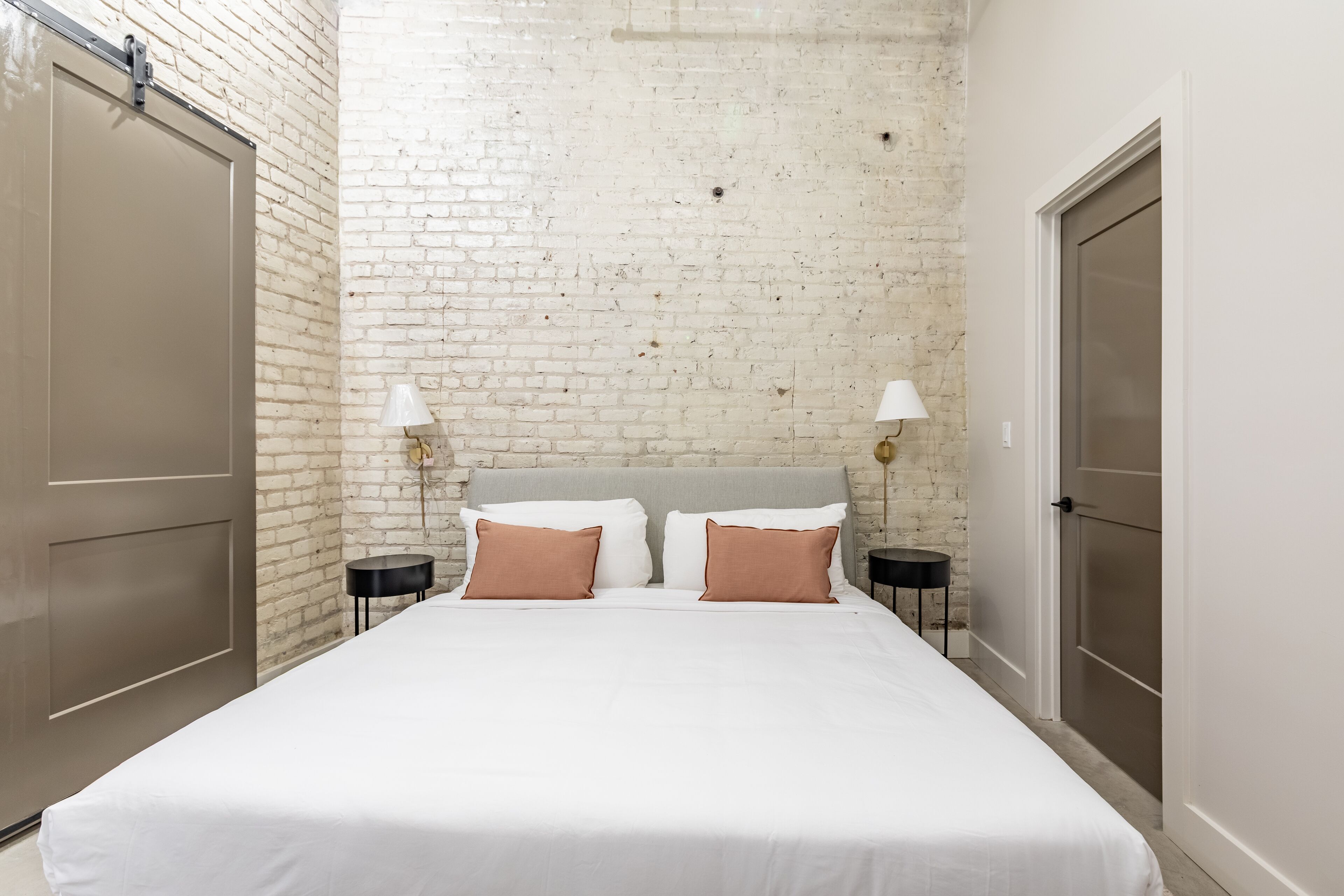 Un dormitorio con pared de ladrillo blanco, una cama con ropa de cama blanca y marrón, dos mesitas de noche con lámparas y una puerta a la derecha.