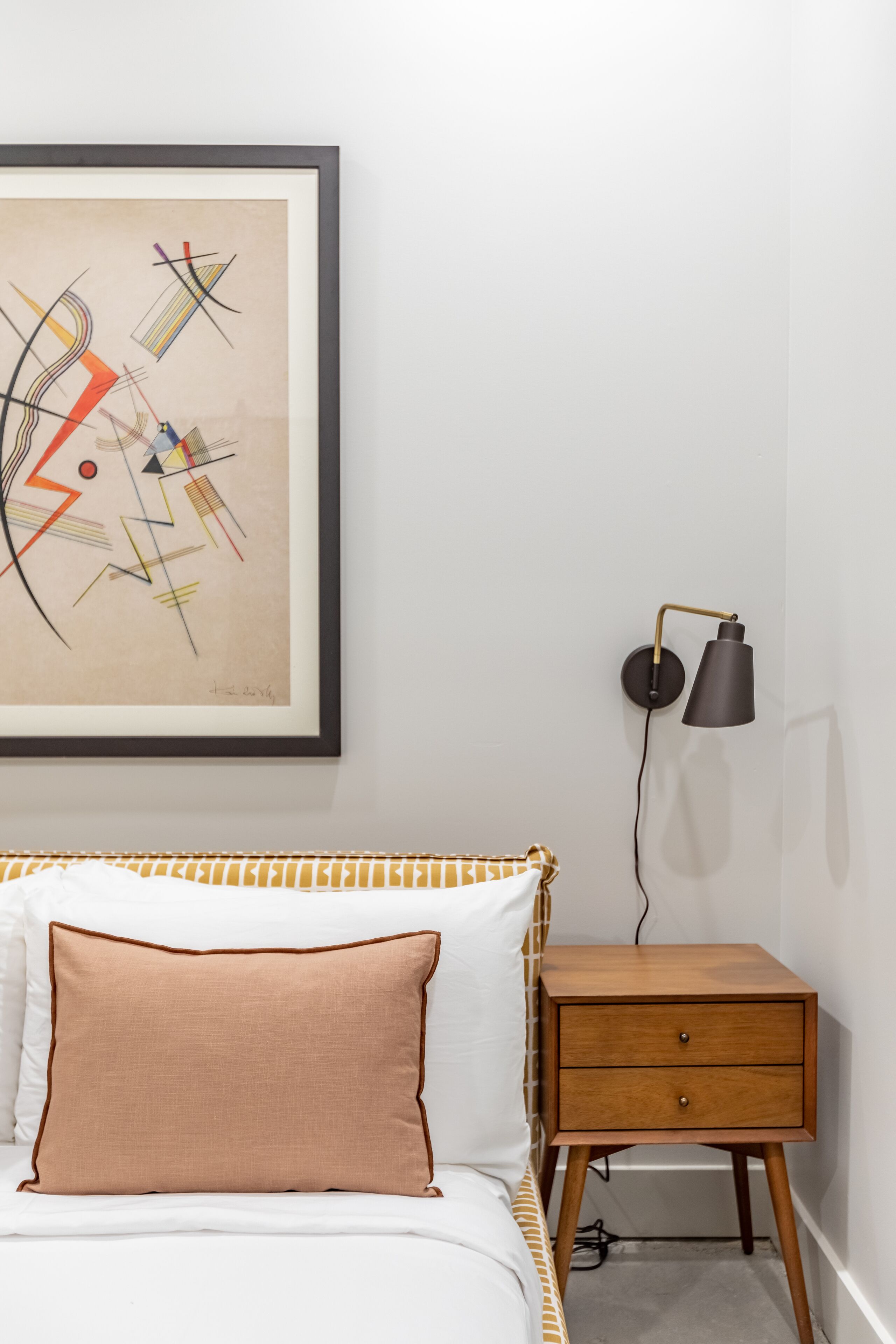 Une chambre à coucher avec un lit, une table de chevet, une lampe et un tableau abstrait encadré accroché au mur.