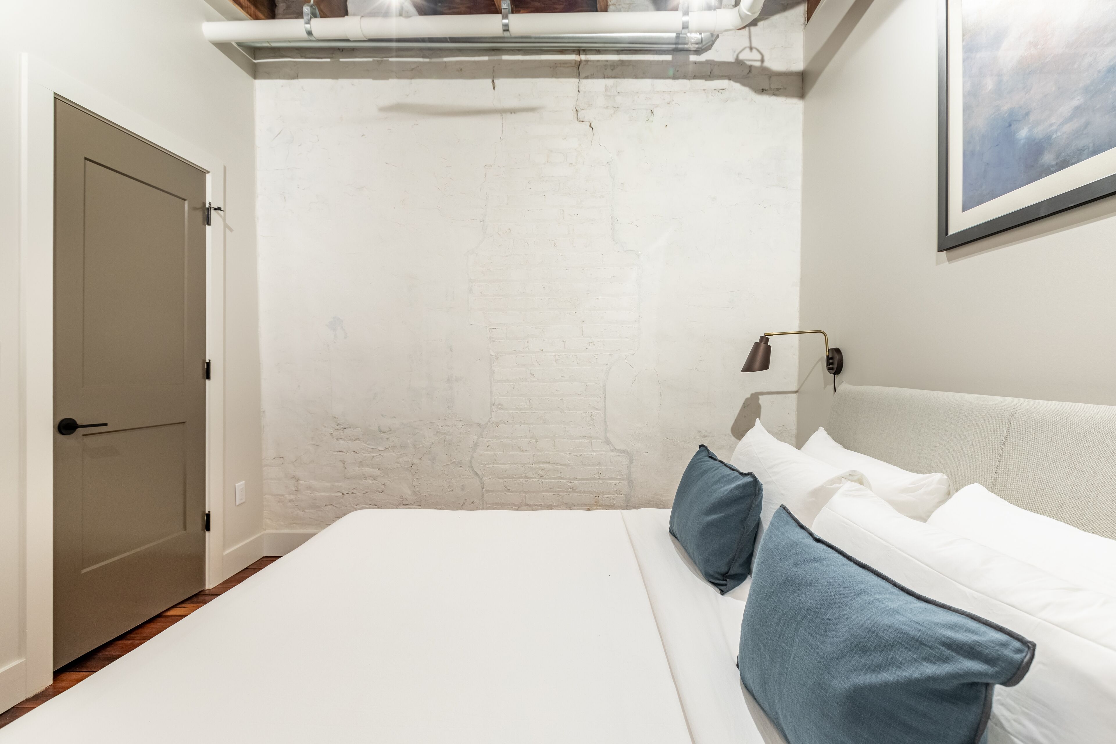 A bedroom with a white brick wall, a bed with white and blue pillows, a bedside table with a lamp, and a door to the left.