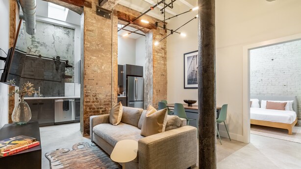 A modern living room with a sofa, a coffee table, and a rug. There is a kitchen in the background, and a bed with a white headboard is visible in the adjacent room. - Ironworks (New Orleans)