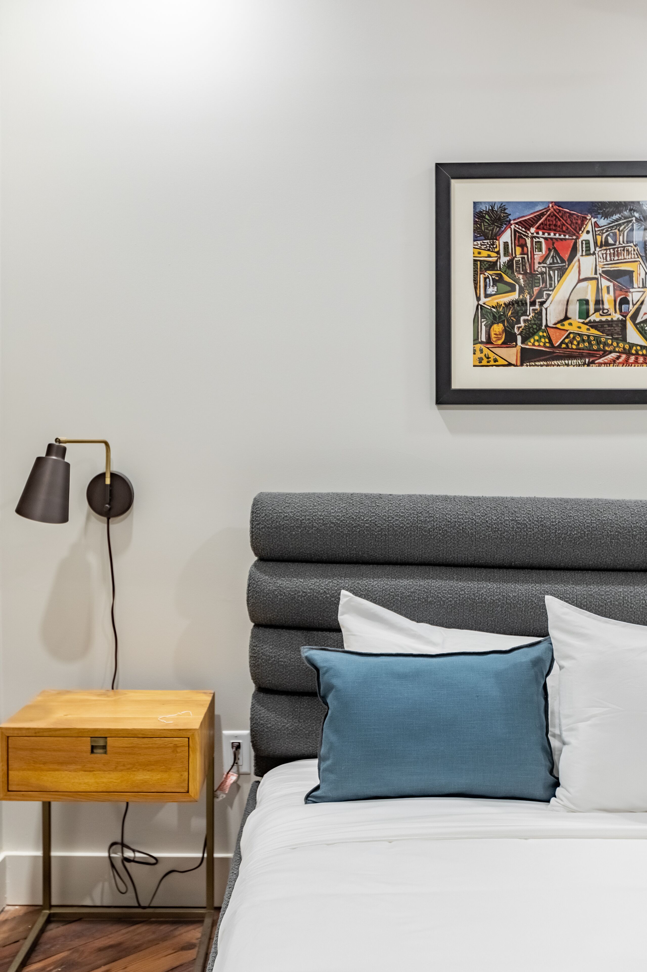 A neatly arranged bedroom with a grey upholstered headboard, a wooden bedside table with a drawer, a framed artwork on the wall, and a lamp beside the table.