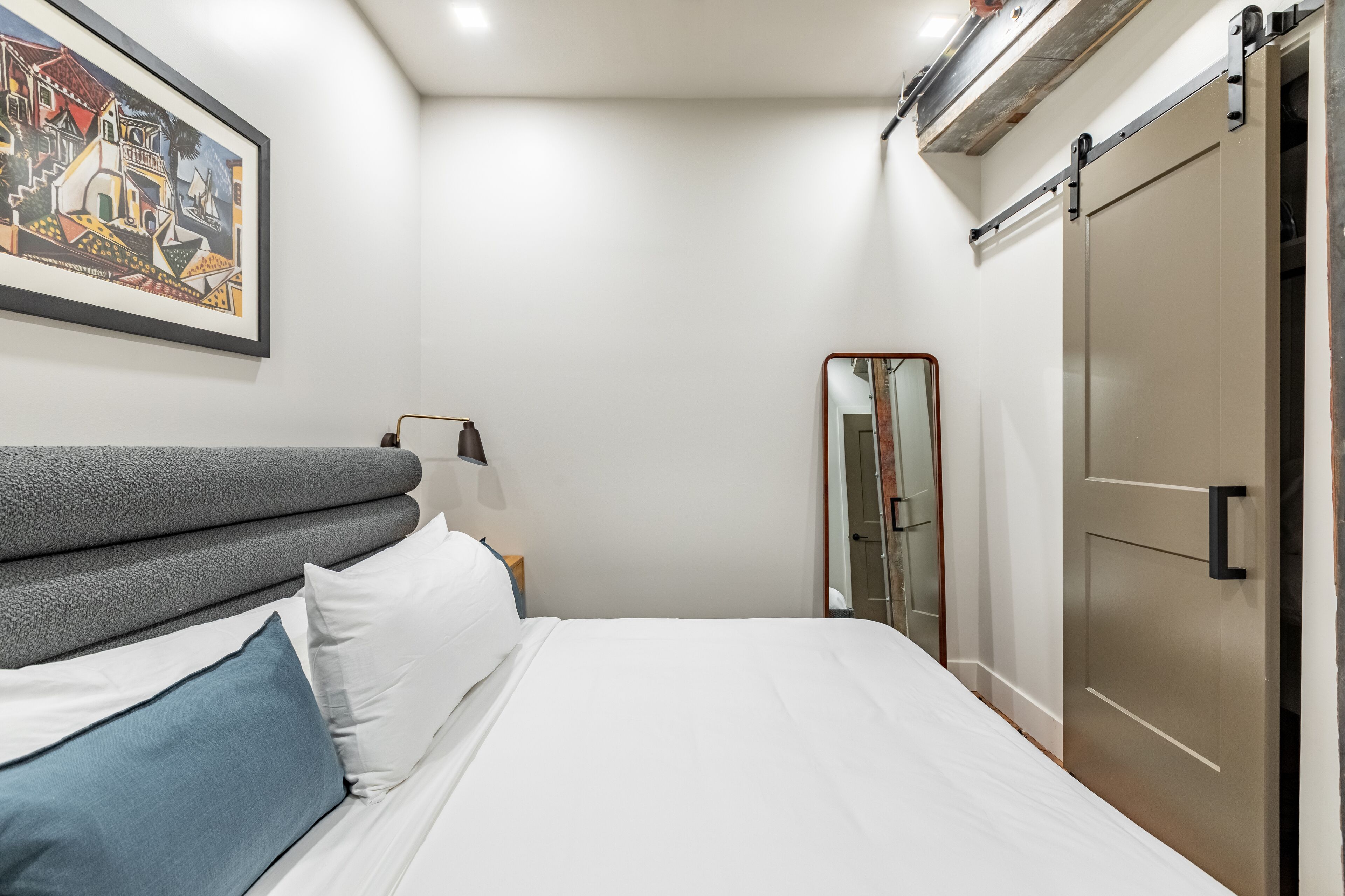A neatly made bed with white and blue pillows, a grey headboard, and a framed artwork on the wall.