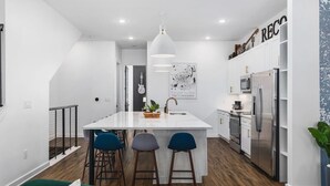 A modern kitchen with white cabinetry, a central island with bar stools, a hanging light fixture, and a framed artwork on the wall.