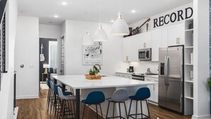 A modern kitchen with white cabinets, a large island with bar stools, a stainless steel refrigerator, and pendant lights.
