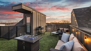 Un patio en la azotea con una parrilla, asientos exteriores y luces colgantes.