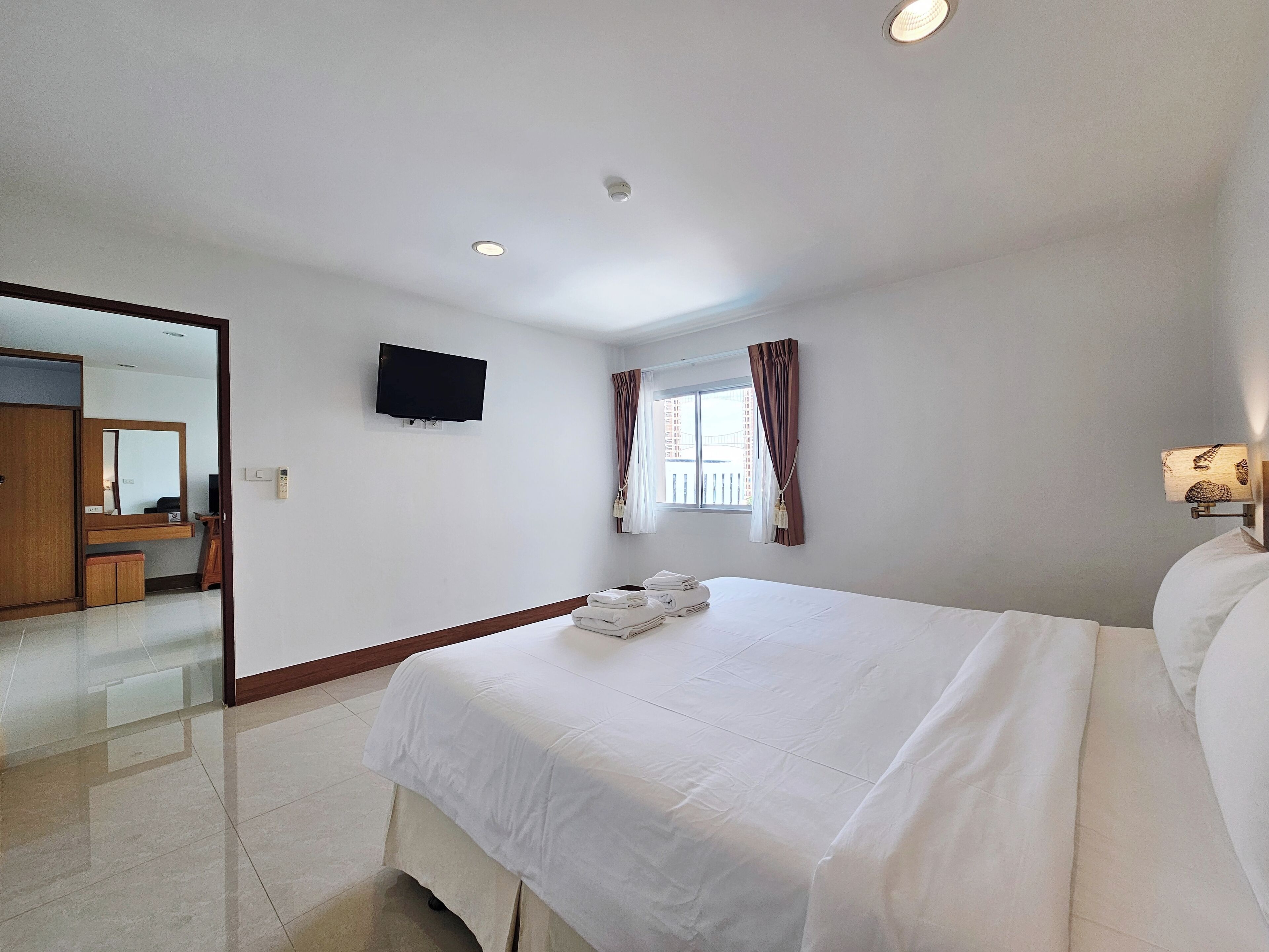 Quarto de hotel com uma cama grande, uma televisão de tela plana na parede, uma janela com cortinas e uma pequena mesa de cabeceira com abajur.