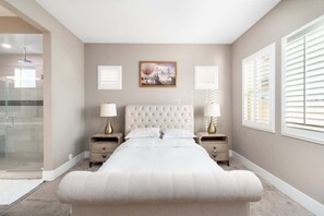 A bedroom with a large bed, two bedside tables, a painting on the wall, and a window with blinds.