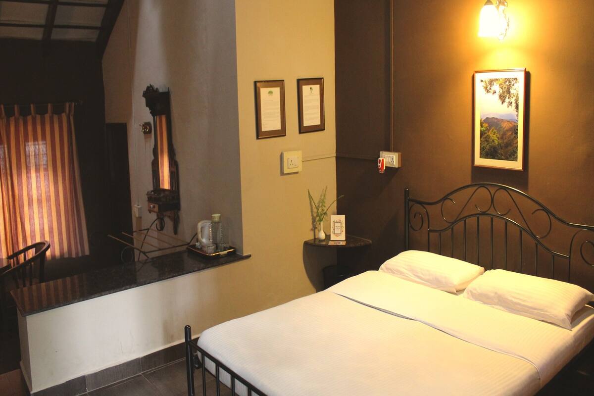 A hotel room with a bed, a desk, a chair, a small table with a plant, and a wall-mounted lamp.
