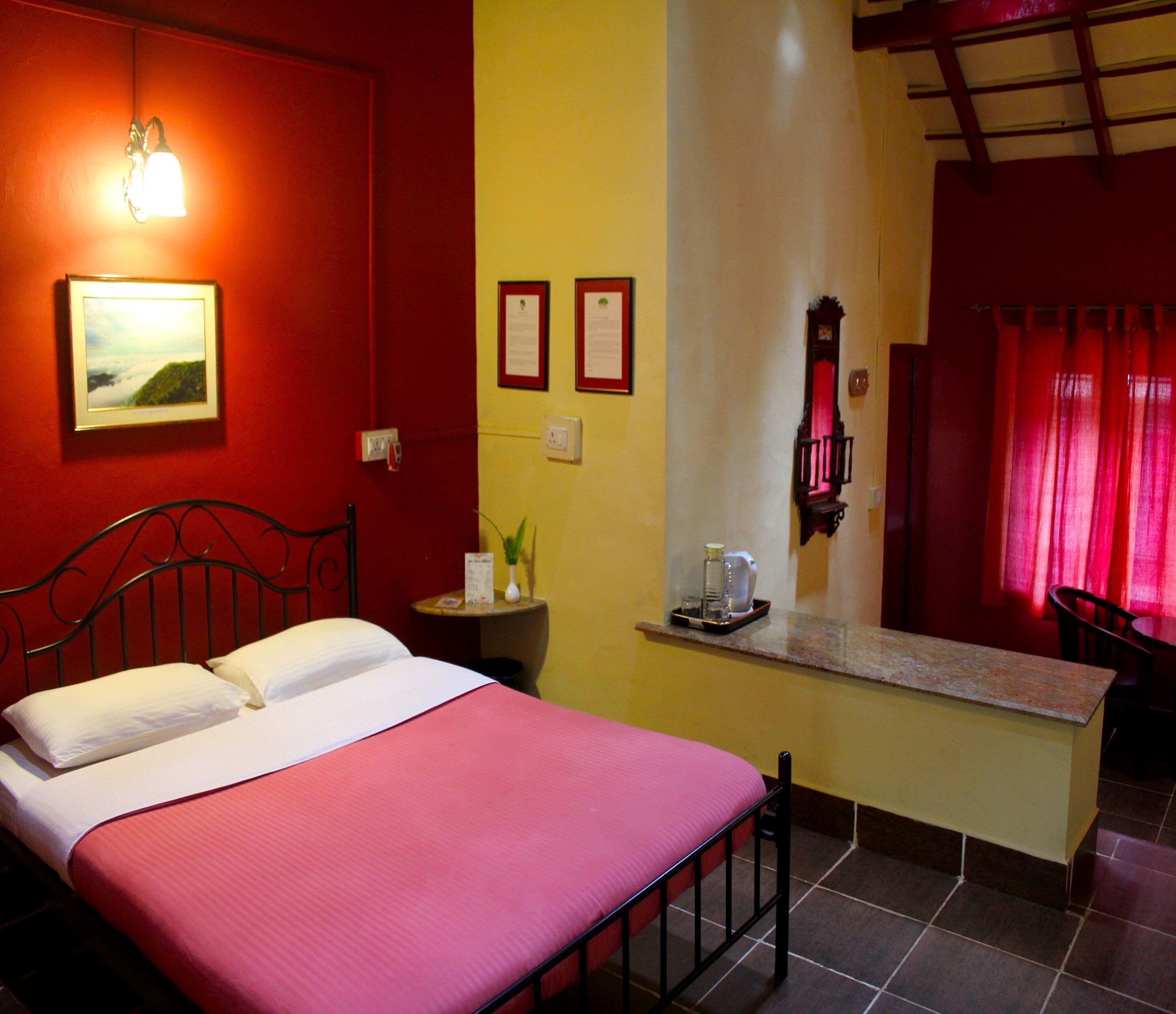 A hotel room with a bed, a small table, a nightstand, a mirror, and a window with red curtains.