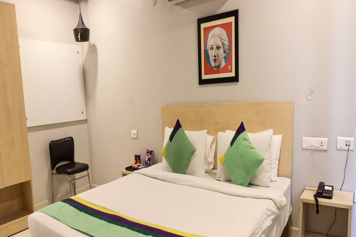Habitación de hotel con cama, una silla, mesita de noche con teléfono y un cuadro en la pared.