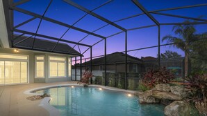 Una piscina interior con techo de cristal, rodeada de rocas y plantas, junto a una casa con amplios ventanales.
