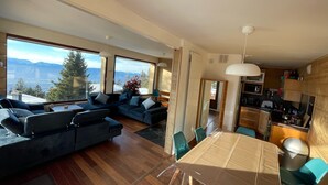 A modern living room with a large sofa, a dining table, and a view of mountains through large windows. - Chalet Chamrousse 1750, 7 Pers, 3 ch, Terrasse, vue Magnifique sur le Vercors (Chamrousse)